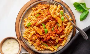 Pyszny makaron z kurczakiem w sosie pomidorowym, udekorowany bazylią. Obok miseczki z parmezanem i ziołami.