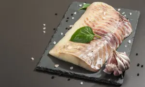 Surowy filet z dorsza czarnego, doprawiony solą i pieprzem, z czosnkiem i listkami bazylii na czarnej desce.