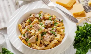 Pyszny makaron z tuńczykiem, pieczarkami i groszkiem w kremowym sosie, udekorowany natką pietruszki. Obok kawałek parmezanu i tarka.