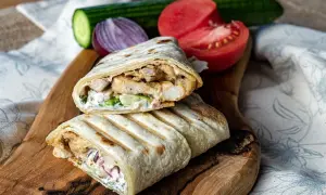 Pyszny kebab w tortilli z kurczakiem, warzywami i sosem, podany na drewnianej desce z pomidorem i ogórkiem.