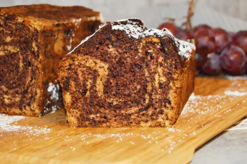 Proste ciasto bananowe z olejem i kakao, z widocznym marmurkowym wzorem. Posypane cukrem pudrem, obok kiść winogron.