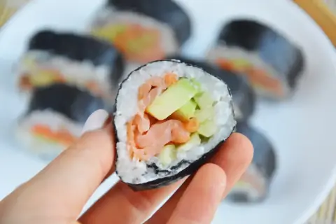 Smaczny futomak z łososiem i awokado, trzymany w dłoni na talerzu pełnym sushi.