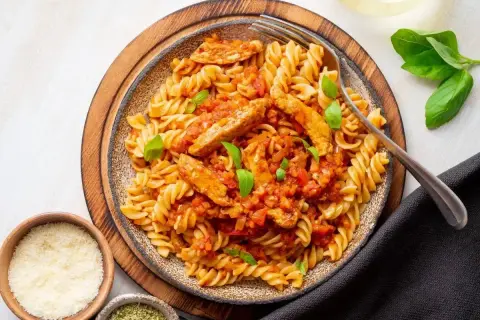 Pyszny makaron z kurczakiem w sosie pomidorowym, udekorowany bazylią. Obok miseczki z parmezanem i ziołami.