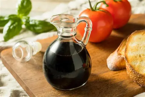 Szklana karafka z ciemnym, gęstym **sosem balsamicznym** obok pomidorów, bazylii i grzanek na desce.