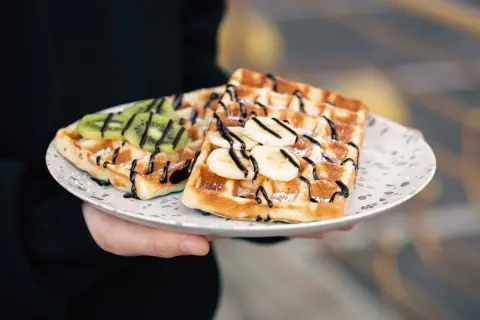 Gofry z bananem i kiwi, polane czekoladą, posypane cukrem perlistym. Pyszny deser na talerzu.