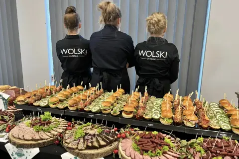 Trzy kobiety w czarnych fartuchach z logo "WOLSKI CATERING" stoją za stołem pełnym przekąsek. To idealna impreza z pysznym jedzeniem.