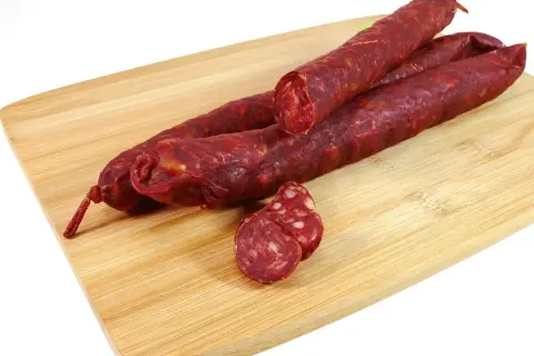 Trzy długie kiełbaski chorizo leżą na drewnianej desce. Jedna jest przekrojona, ukazując charakterystyczne kawałki mięsa i tłuszczu.