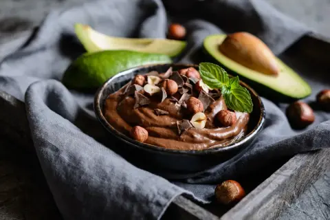 Kremowa nutella z awokado, udekorowana orzechami laskowymi, kawałkami czekolady i miętą. Obok leżą połówki awokado.