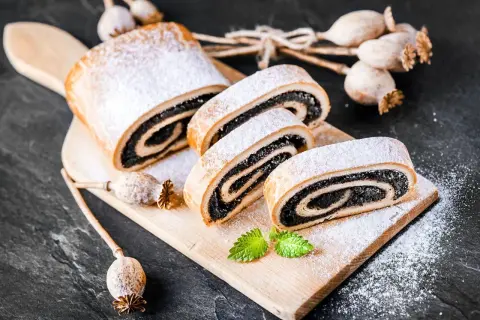 Stary przepis na makowiec, pyszny, zawinięty w ciasto i posypany cukrem pudrem. Obok suszone maki.