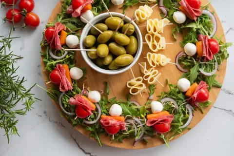 Kolorowe finger food: szaszłyki z mozzarellą, pomidorkami, salami i warzywami, oliwki i serowe zawijasy na desce.