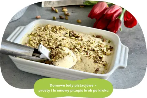 Domowe lody pistacjowe w białej misce, posypane orzechami. Obok czerwone tulipany.