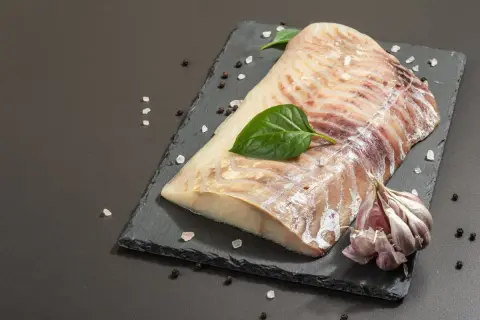 Surowy filet z dorsza czarnego, doprawiony solą i pieprzem, z czosnkiem i listkami bazylii na czarnej desce.
