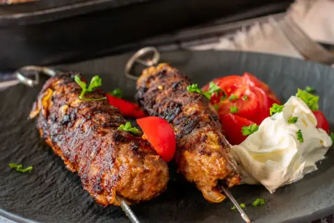 Dwa soczyste kofta kebab na patyczkach, podane z pomidorami i kremowym sosem.