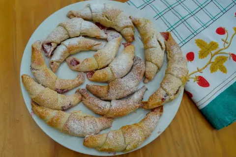 Kruche rogaliki z marmoladą, posypane cukrem pudrem, ułożone na talerzu.