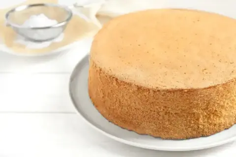 Idealny biszkopt, by wyszedł puszysty, piecz w temperaturze 170-180°C. Na talerzu leży złocisty biszkopt, obok sitko z cukrem pudrem.