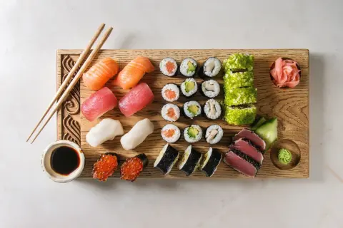 Drewniana deska z różnorodnymi składnikami do sushi: nigiri z łososiem, tuńczykiem i krewetkami, rolki z awokado i kurczakiem, maki, ikra, sos sojowy, wasabi i marynowany imbir.