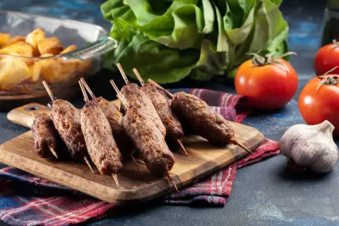 Aksamitne ćevapi na drewnianej desce, obok pomidory, czosnek i pieczone ziemniaki.