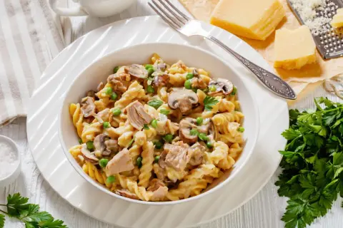 Pyszny makaron z tuńczykiem, pieczarkami i groszkiem w kremowym sosie, udekorowany natką pietruszki. Obok kawałek parmezanu i tarka.