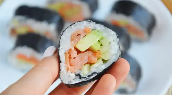 Smaczny futomak z łososiem i awokado, trzymany w dłoni na talerzu pełnym sushi.