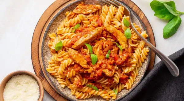 Pyszny makaron z kurczakiem w sosie pomidorowym, udekorowany bazylią. Obok miseczki z parmezanem i ziołami.