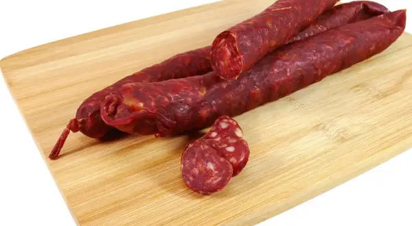 Trzy długie kiełbaski chorizo leżą na drewnianej desce. Jedna jest przekrojona, ukazując charakterystyczne kawałki mięsa i tłuszczu.