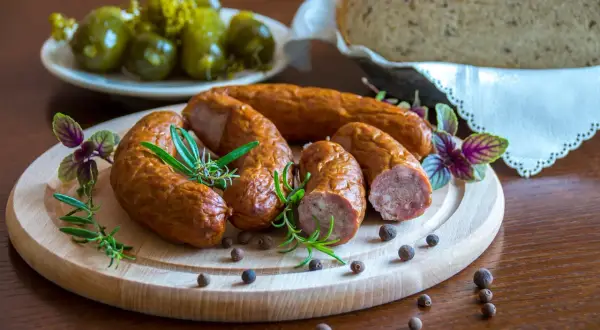 Kiełbasa podwawelska na drewnianej desce z rozmarynem i pieprzem. W tle ogórki i chleb.
