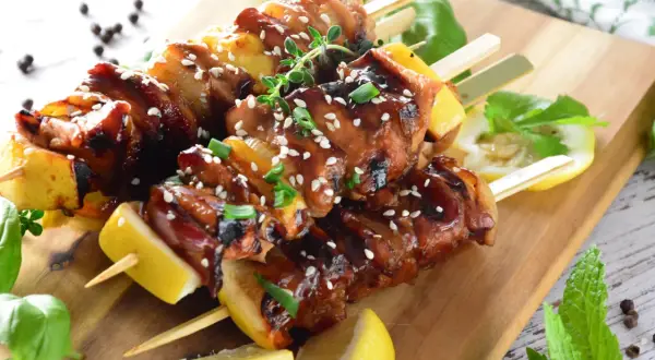 Aromatyczne yakitori z kurczaka w sosie teriyaki, posypane sezamem i szczypiorkiem, podane z cytryną na drewnianej desce.