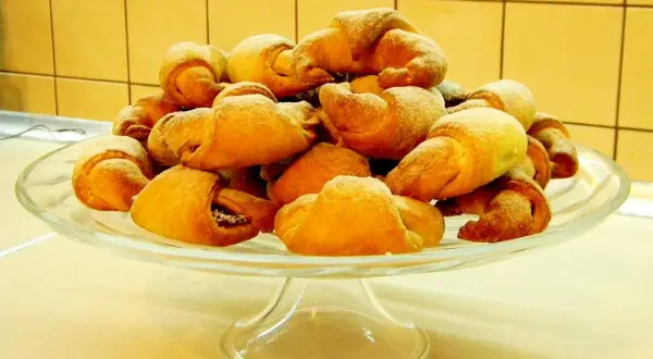 Rogaliki drożdżowe z przepisu babci, posypane cukrem pudrem, piętrzą się na szklanej paterze.