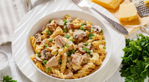Pyszny makaron z tuńczykiem, pieczarkami i groszkiem w kremowym sosie, udekorowany natką pietruszki. Obok kawałek parmezanu i tarka.