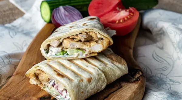 Pyszny kebab w tortilli z kurczakiem, warzywami i sosem, podany na drewnianej desce z pomidorem i ogórkiem.