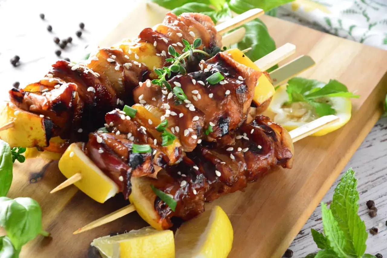 Aromatyczne yakitori z kurczaka w sosie teriyaki, posypane sezamem i szczypiorkiem, podane z cytryną na drewnianej desce.