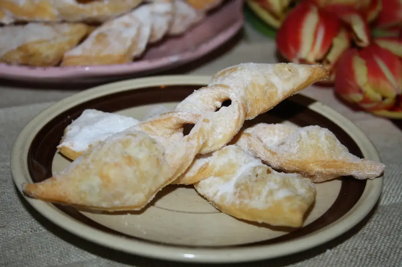 Chrupiące faworki, jak z przepisu babci, posypane cukrem pudrem, leżą na talerzu.