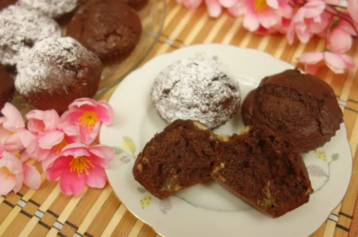 Puszyste ciasto bananowe na oleju, czekoladowe muffinki posypane cukrem pudrem, obok różowe kwiaty.