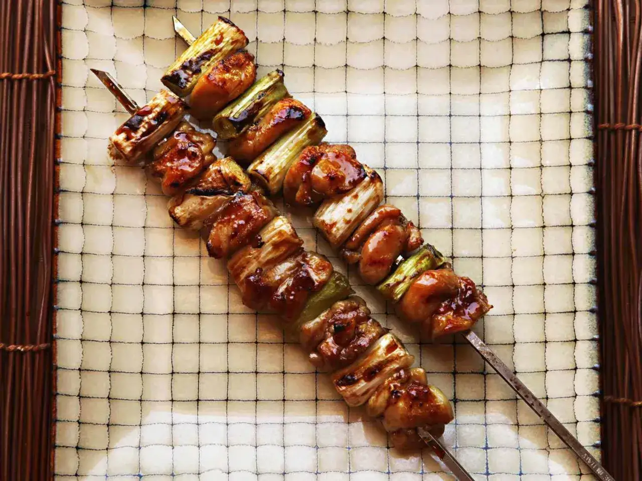 Dwa apetyczne szaszłyki yakitori z kurczakiem i zieloną cebulką, polane słodkim sosem, leżą na talerzu z siatką.