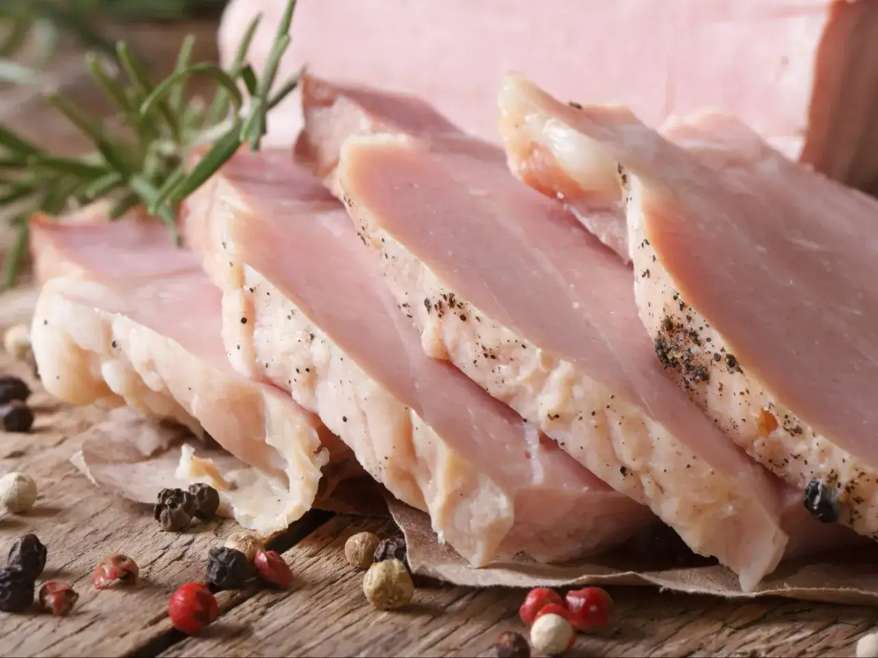 Pokrojony schab gotowany 2 razy po 10 minut, z gałązką rozmarynu i ziarnami pieprzu na drewnianym blacie.