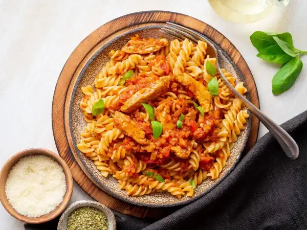 Pyszny makaron z kurczakiem w sosie pomidorowym, udekorowany bazylią. Obok miseczki z parmezanem i ziołami.