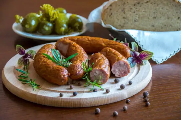 Kiełbasa podwawelska na drewnianej desce z rozmarynem i pieprzem. W tle ogórki i chleb.